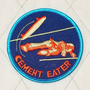 May include: A blue and orange embroidered patch with a cartoon figure falling off a skateboard. The text "Cement Eater" is written in orange around the edge of the patch.