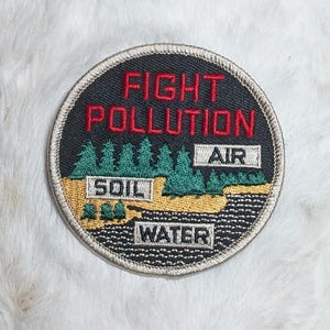 May include: A circular embroidered patch with a black background featuring the red text FIGHT POLLUTION. The design includes green pine trees, a tan land area labeled SOIL, a white box labeled AIR, and a black water area labeled WATER.