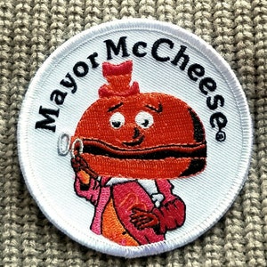May include: White embroidered patch with a cartoon character wearing a red and black hat and a red and black suit. The character has a large red and black burger for a head. The text "Mayor McCheese" is written in a circle around the character.