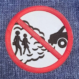May include: A circular embroidered patch featuring a white background with a red border and a red diagonal slash. The design depicts a black silhouette of a car emitting smoke toward two black silhouettes of people walking, all enclosed by the red border.