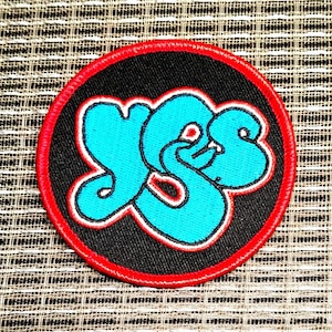 May include: A circular embroidered patch featuring a black background with a bright red stitched border. In the center, the word YES is displayed in a stylized, bubble-letter font in bright cyan blue with a thin white and red outline.
