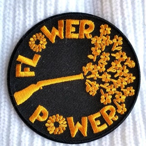 May include: A round black patch with the words "FLOWER POWER" in yellow, surrounding a yellow rifle that appears to be shooting a spray of yellow flowers. The patch is on a white, textured surface.