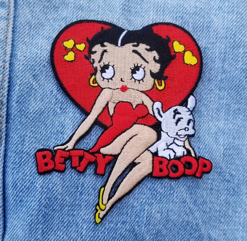 May include: An embroidered patch featuring the cartoon character Betty Boop with black hair, a red dress, and gold hoop earrings, positioned in front of a large red heart with yellow hearts. A small white dog sits beside her with the text BETTY BOOP in red.