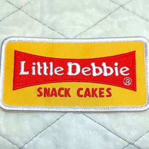 May include: A yellow and red embroidered patch with the text "Little Debbie Snack Cakes" and a registered trademark symbol.
