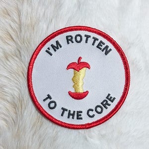 May include: A circular embroidered patch with a white background and a red border. The center features a yellow apple core with red top and bottom sections. The black text reads I'M ROTTEN TO THE CORE, arched around the apple core illustration.