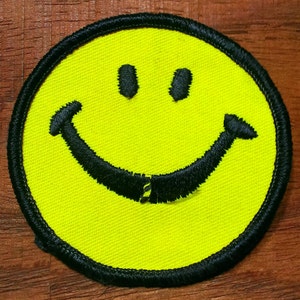 May include: A yellow smiley face patch with a black outline and a black stitched smile. The patch is made of fabric and has a rough texture.