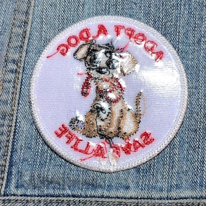 May include: A circular embroidered patch featuring a tan puppy with a red collar and leash. The text "ADOPT A DOG" is arched across the top and "SAVE A LIFE" is arched across the bottom in red thread on a light lavender background.