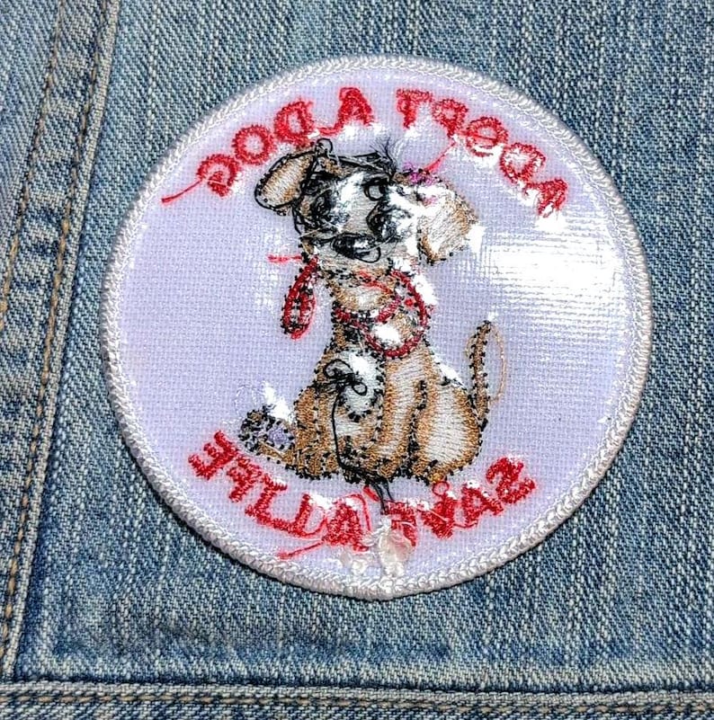 May include: A circular embroidered patch featuring a tan puppy with a red collar and leash. The text "ADOPT A DOG" is arched across the top and "SAVE A LIFE" is arched across the bottom in red thread on a light lavender background.
