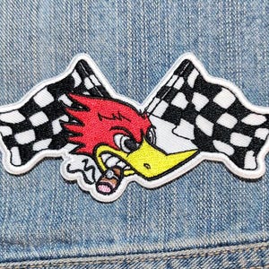 May include: An embroidered patch featuring a red cartoon bird with a yellow beak, smoking a brown cigar, positioned in front of two crossed black and white checkered racing flags. The patch has a white border and is placed on blue denim fabric.