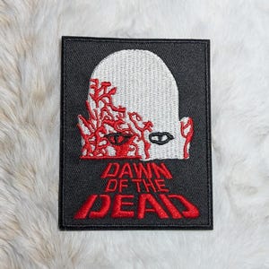May include: A rectangular black embroidered patch featuring a white head silhouette with red vein-like details and a single black eye. Below the graphic, the text DAWN OF THE DEAD is stitched in bold red capital letters with a black outline.