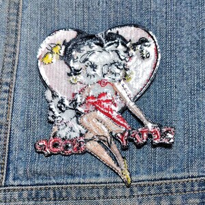 May include: An embroidered patch featuring a cartoon character with black hair and a red dress centered inside a heart shape. The character is positioned above red embroidered text that reads "BOOP-OOP-A-DOOP" against a light pink background.