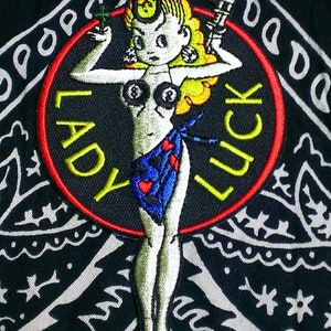 May include: A red, white, and yellow embroidered patch with a cartoon pinup girl holding a pair of dice and a gun. The patch says "Lady Luck" in a yellow circle.