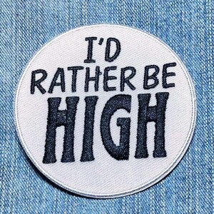 May include: A circular embroidered patch with a white background and a dark blue border. The patch features the black embroidered text I'D RATHER BE HIGH centered in the middle, with the word HIGH appearing in a larger font size than the others.