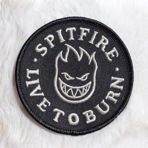 May include: Round black embroidered patch with the words "SPITFIRE" and "LIVE TO BURN" in white. The patch features a white graphic of a flaming skull with sharp teeth.
