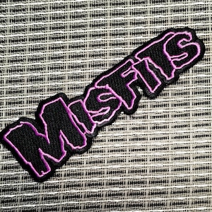 May include: Black embroidered patch with the word "MISFITS" in a stylized font. The text is outlined in purple and has a black background. The patch is likely for clothing or accessories.