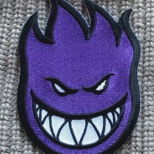 May include: Purple and black embroidered patch with a cartoon face with a wide, toothy grin. The face is set against a flame-like background.