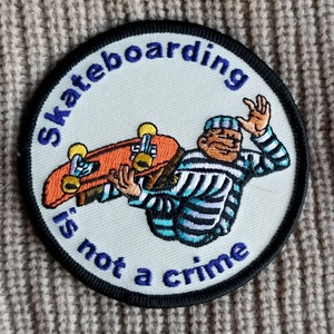 May include: A black and white embroidered patch with a cartoon of a person in a striped shirt skateboarding. The text "Skateboarding is not a crime" is written in a circle around the image.