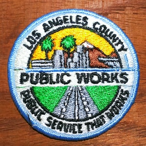 May include: A circular embroidered patch featuring the text LOS ANGELES COUNTY PUBLIC WORKS and PUBLIC SERVICE THAT WORKS. The design includes a yellow sky, green palm trees, a mountain, a gray cityscape, and a gray road leading into the distance.