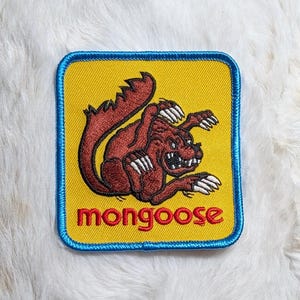 May include: A square embroidered patch with a yellow background and a blue border. It features a brown mongoose illustration with white claws and teeth, positioned above the red lowercase text "mongoose" centered at the bottom of the patch.