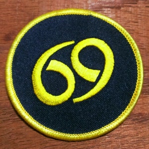 May include: A round, black patch with a yellow embroidered number "69" in the center. The patch has a yellow border.
