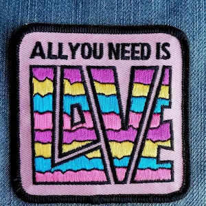 May include: Pink fabric patch with black stitching and the text "All you need is" above a colorful, striped, embroidered word "LOVE".