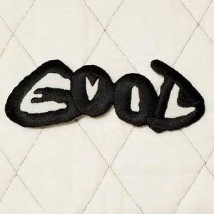 May include: Embroidered patch with the word "GOOD" in black, outlined in white, on a white quilted background. The patch has a bold, stylized font.