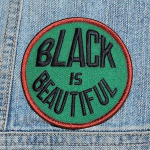 May include: A circular embroidered patch featuring a vibrant green background with a dark red border. The text "BLACK IS BEAUTIFUL" is stitched in bold black lettering across the center of the patch, arranged in a curved, stacked layout.