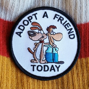 May include: White embroidered patch with black stitching. The patch is round and features two cartoon dogs with the text "Adopt a Friend Today".