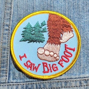 May include: A circular embroidered patch with a light blue background, a yellow border, and a brown hairy foot. It features green pine trees and the red text I SAW BIG FOOT. The patch is attached to a blue denim fabric surface with visible stitching.