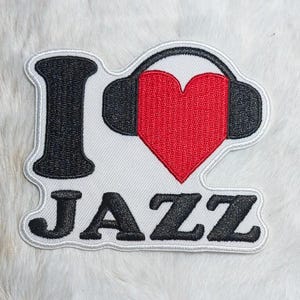 May include: An embroidered patch featuring the text I, a red heart shape positioned inside black headphones, and the text JAZZ below. The design is rendered in black, red, and white thread with a white border, creating a clear graphic emblem.