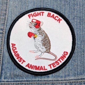 May include: A circular embroidered patch with a white background and a black border. It features a brown and white mouse wearing a red boxing helmet and red boxing gloves. The red text reads FIGHT BACK AGAINST ANIMAL TESTING around the edge.
