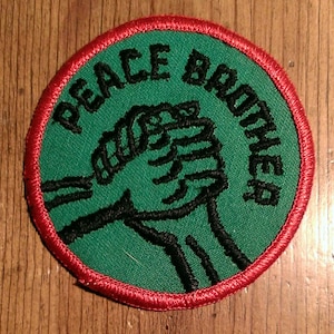 May include: Green embroidered patch with red stitching around the edge. The patch features two clasped hands and the text "Peace Brothers".