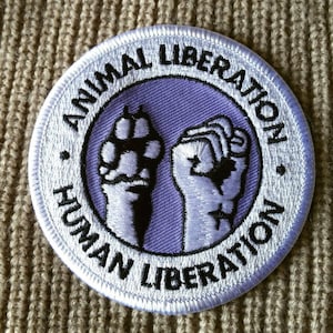 May include: White embroidered patch with the text "Animal Liberation - Human Liberation" in black lettering around a purple circle with two fists raised in the air, one with a paw print.