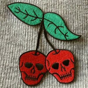 May include: Two red cherries with skull faces and green leaves. The cherries are embroidered on a gray fabric.