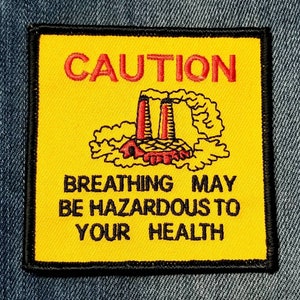 May include: Yellow caution sign with black border, featuring a red and black illustration of a factory emitting smoke. The text reads "CAUTION" in red and "BREATHING MAY BE HAZARDOUS TO YOUR HEALTH" in black.