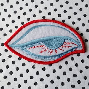 May include: A red and blue embroidered eye patch with a white background. The eye is closed and has lightning bolts coming out of it.
