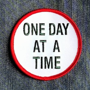 May include: A circular patch with a white center and a red border. The patch features the embroidered text "ONE DAY AT A TIME" in dark green letters. The patch is likely designed to be sewn or ironed onto clothing or accessories.