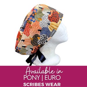 May include: A scrub cap featuring a colorful pumpkin pattern with various textures and spots in shades of orange, blue, green, and black. The cap has a black ribbon tie at the back and a small grey tag with the text SCRIBES printed in white.
