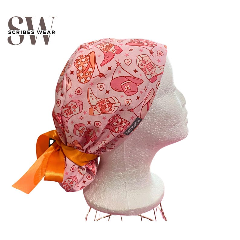 May include: Pink surgical scrub hat with a tie-back design. The hat features a pattern of pink cowboy boots, hats, and stars. The tie-back is orange satin.