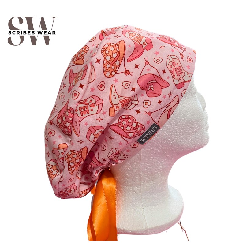 May include: Pink surgical scrub hat with a cowboy boot and hat print. The hat has a bright orange ribbon tie.