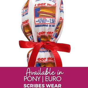 May include: A white scrub cap featuring a repeating graphic print of a hot dog and the text I GOT THAT DOG IN ME. The design includes menu-style details like 1.50 and 580-850 cal. A bright red satin ribbon is tied in a bow around the center.