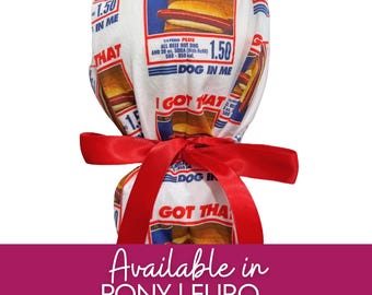 May include: A white scrub cap featuring a repeating graphic print of a hot dog and soda combo with the text I GOT THAT DOG IN ME, 1.50, and 580-850 cal. The cap is tied with a bright red satin ribbon at the back, securing the fabric in a gathered style.