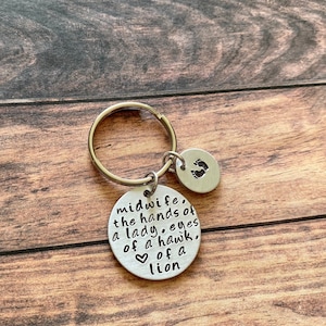May include: Silver keyring with a round charm that reads "midwife, the hands of a lady, eyes of a hawk, ♡ of a lion". A smaller charm with a footprint design is attached to the keyring.
