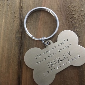 May include: Silver dog bone-shaped keyring with a ring. The bone has the text "i'm why we can't have nice things" and "DUDLEY 777.333.9999" engraved on it.