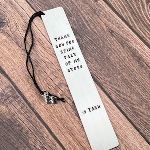 May include: Silver bookmark with the text "Thank you for being part of my story" and a heart with the name "Tash" below. A small silver charm shaped like a book is attached to the bookmark.