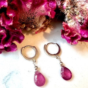 May include: A pair of silver hoop earrings with a dangling teardrop-shaped pink gemstone. The earrings are on a white surface.
