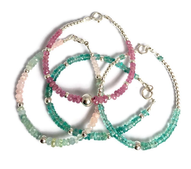 May include: Three silver beaded bracelets with pink, green, and light blue gemstones. The bracelets are layered on top of each other.