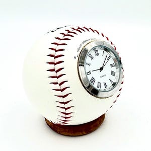 May include: A baseball-shaped clock with a white exterior and red stitching. The clock face has Roman numerals and a silver bezel. The clock sits on a small, round, wooden base. The text "SOLID CORK & RUBBER" is visible on the baseball.