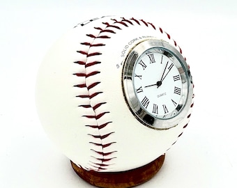 May include: A baseball-shaped clock with a white exterior and red stitching. The clock face has Roman numerals and a silver bezel. The clock sits on a small, round, wooden base. The text "SOLID CORK & RUBBER" is visible on the baseball.