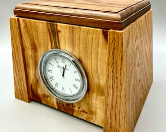 May include: A wooden box with a clock face on the front. The clock face is white with black Roman numerals and black hands. The box is made of light brown wood with a darker brown top.